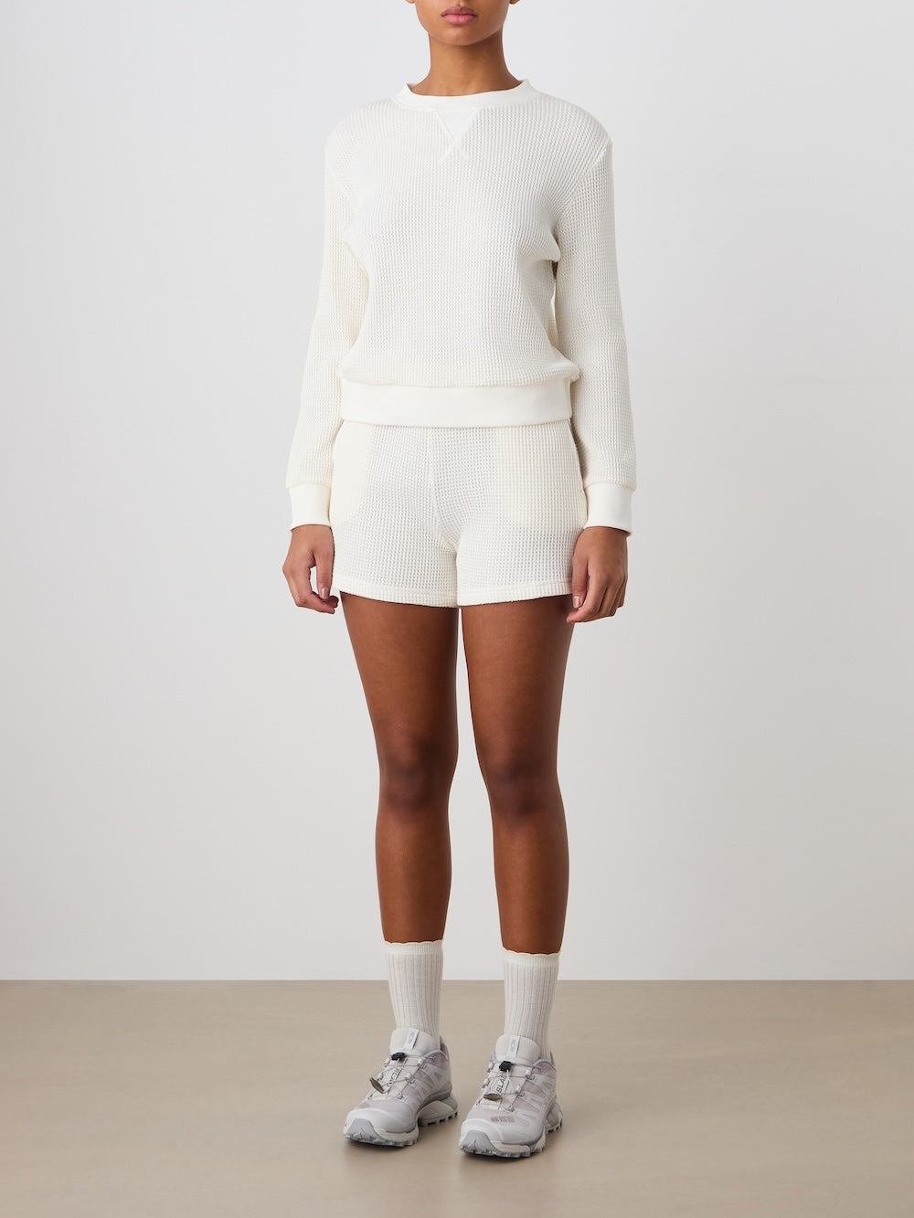A person wears the We Norwegians Waffle Crewneck Women sweater in cream with matching shorts, light gray crew socks, and gray sneakers. Styled in a relaxed fit against a plain background, face not visible.