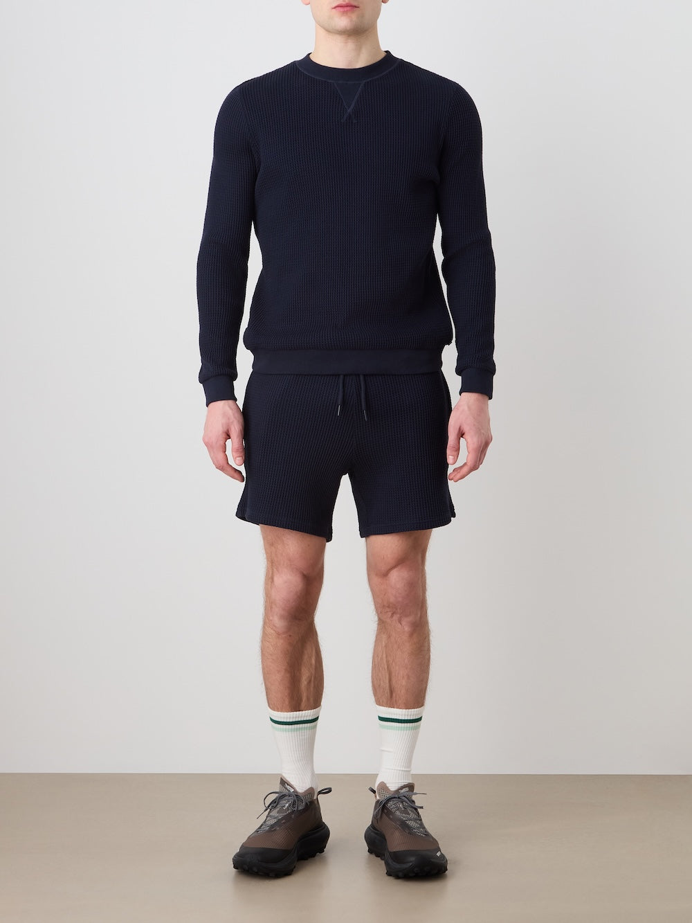 A man faces forward against a plain light background, wearing the We Norwegians Waffle Crewneck Men sweater in navy, matching navy shorts, white socks with green stripes, and black sneakers. His arms are relaxed at his sides.