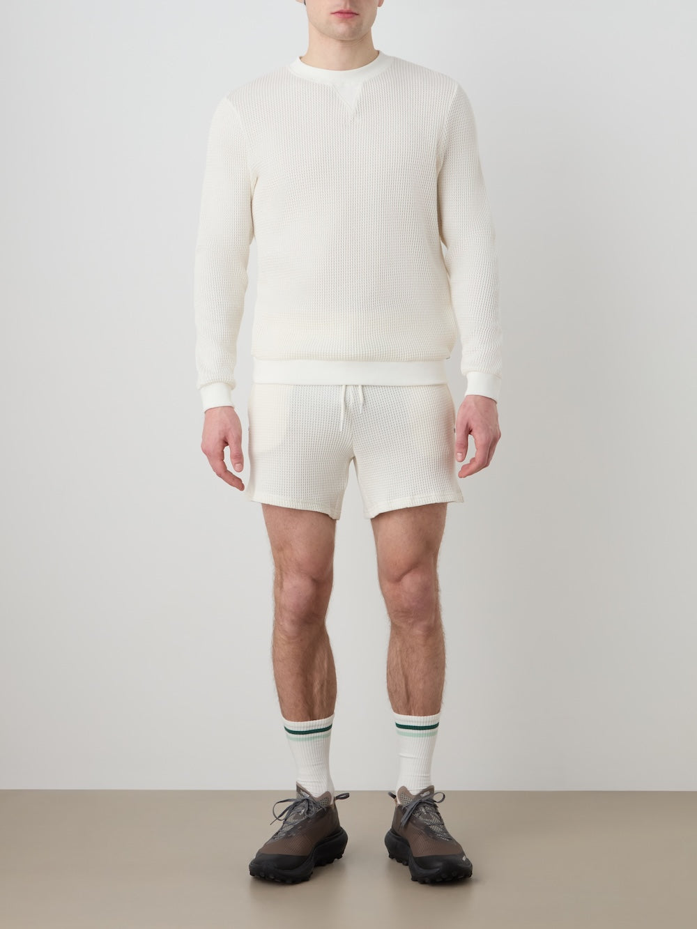 A man, partially out of frame, stands against a plain background wearing the We Norwegians Waffle Crewneck Men shirt with matching shorts, white socks with green stripes, and black sneakers.
