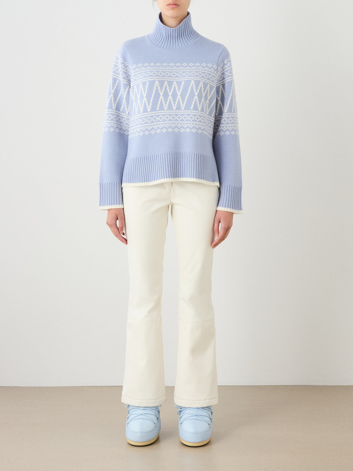 A person wearing the Setesdal Pullover women sweater in light blue by We Norwegians, paired with cream-colored pants and light blue boots, stands against a plain background facing forward with their hands by their sides.