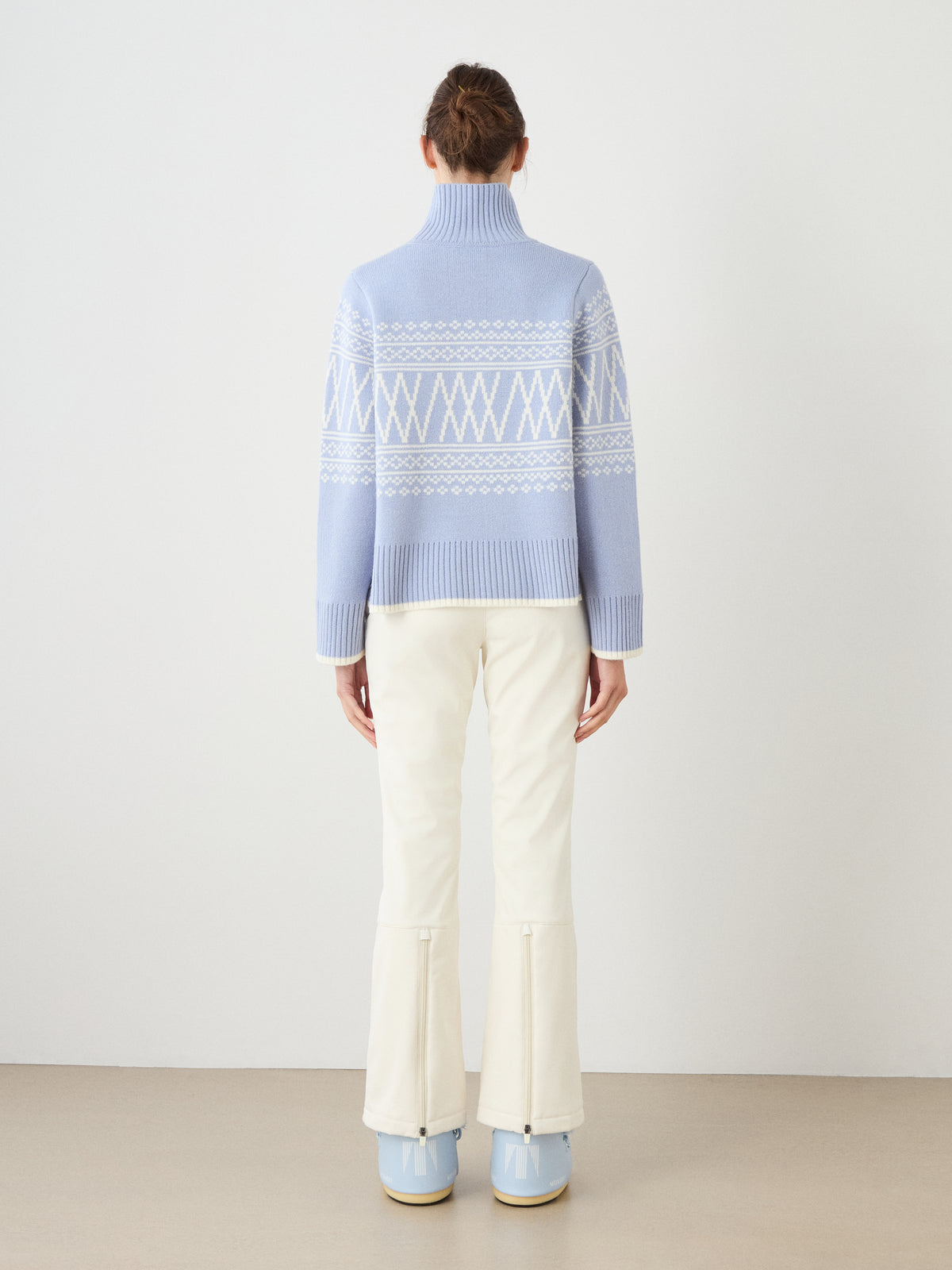 A person stands facing away, wearing a We Norwegians Setesdal Pullover women’s turtleneck sweater in a light blue and white pattern, paired with off-white pants featuring back hem zippers and light blue boots, set against a plain white wall and beige floor.