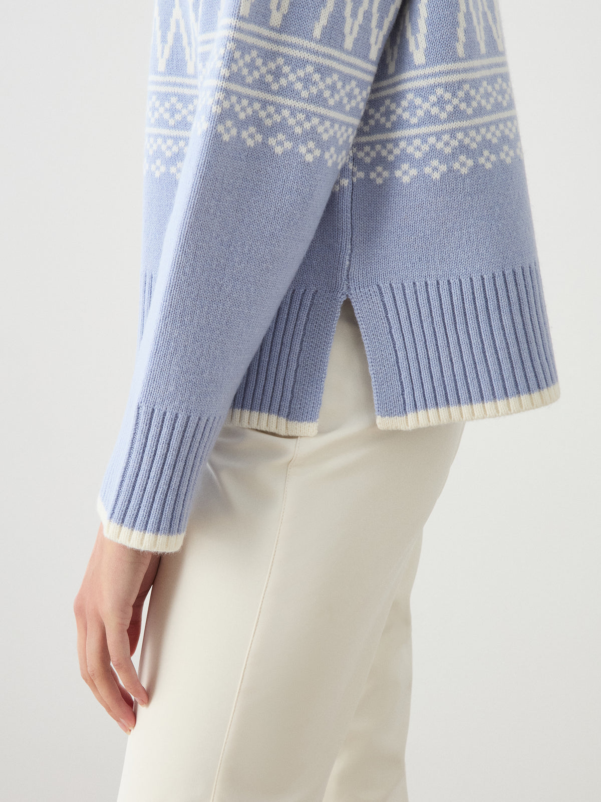 A person wearing the We Norwegians Setesdal Pullover women in light blue, featuring classic white Norwegian knit patterns, paired with beige pants. This merino-cashmere sweater includes ribbed cuffs and hem as well as a side slit.