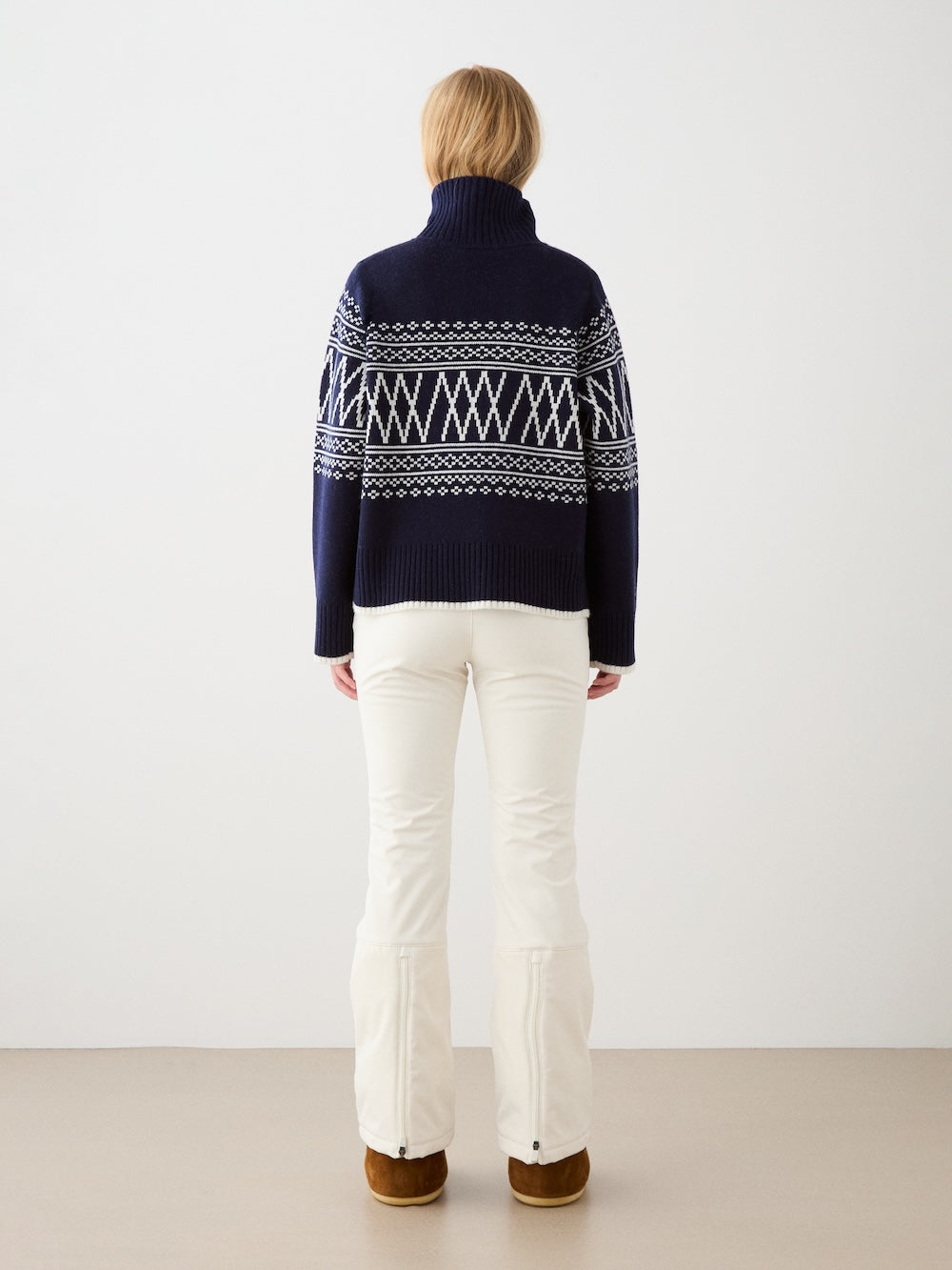 A woman with light brown hair wears the We Norwegians Setesdal Pullover, featuring a navy and white Norwegian pattern, paired with white pants and brown shoes. She stands indoors facing away from the camera against a plain background.