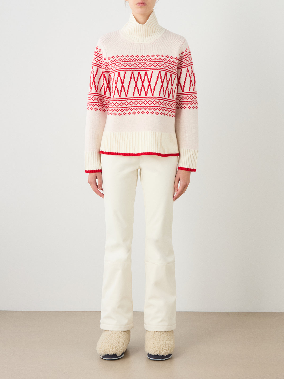A person wearing the We Norwegians Setesdal Pullover women’s sweater with classic red Norwegian knit patterns, paired with cream-colored pants and beige fuzzy boots, stands against a plain background.