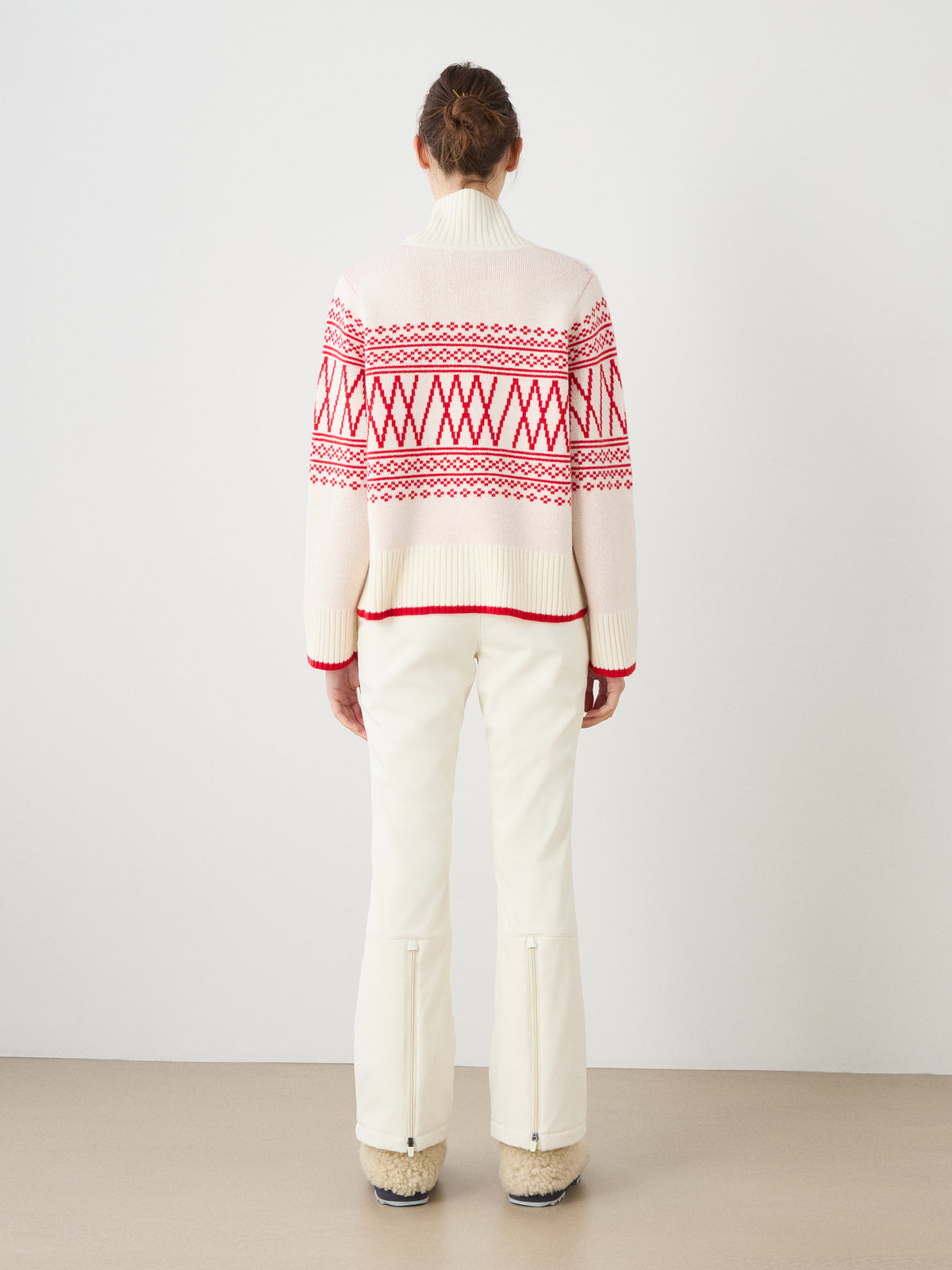 A person stands facing away in a minimal indoor setting with a plain light-colored wall and floor, wearing the We Norwegians Setesdal Pullover women in white with red geometric patterns, paired with white pants and beige shoes.