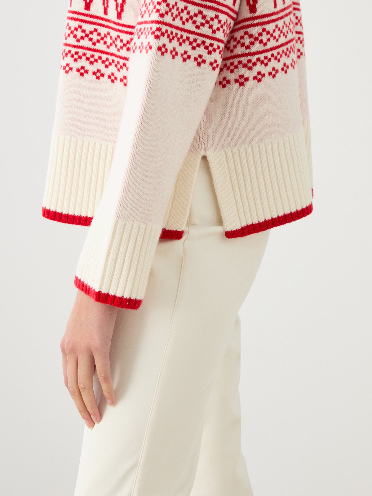 A woman wearing off-white pants and the We Norwegians Setesdal Pullover, featuring cream fabric with red Norwegian knit patterns and ribbed cuffs, stands against a plain background.