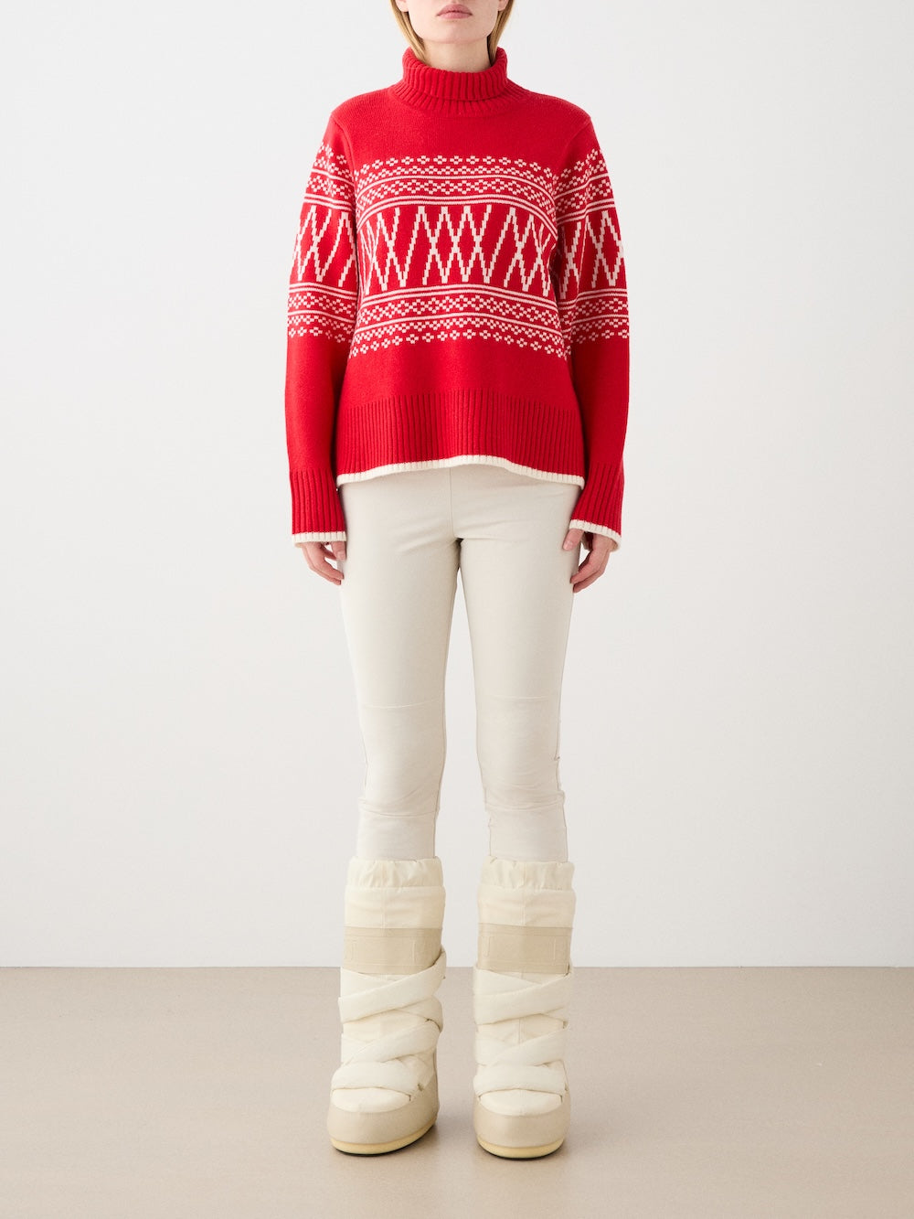 A woman models the We Norwegians Setesdal Pullover in red and white, paired with white pants and puffy winter boots. She stands against a plain background, face turned away, with arms relaxed at her sides.