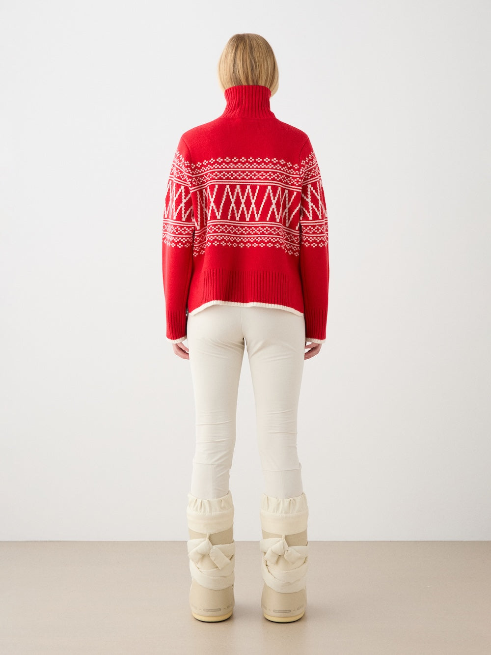 A person stands facing away, wearing the We Norwegians Setesdal Pullover women in red and white, paired with off-white pants and beige padded winter boots against a plain light background.