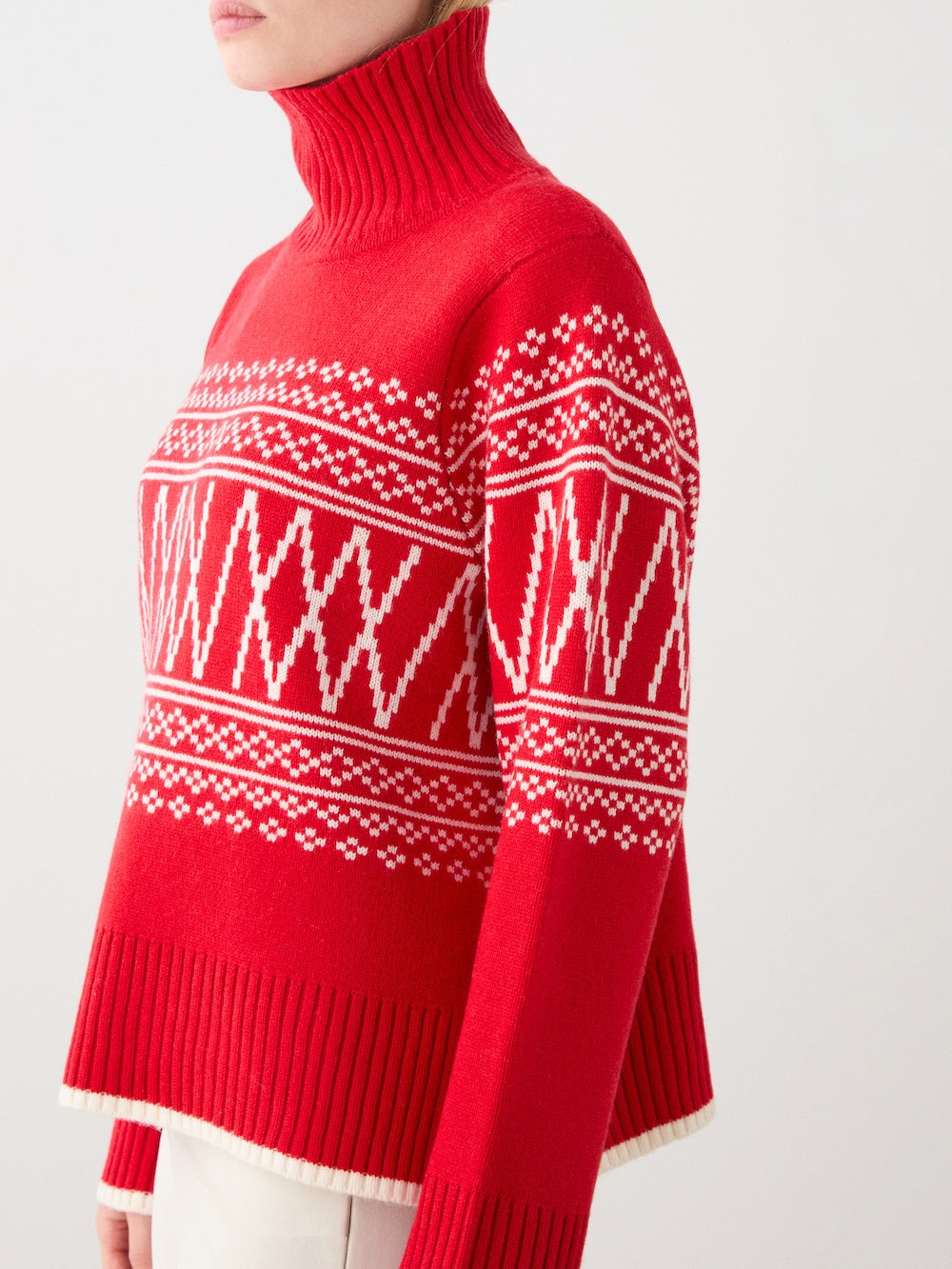 A woman is seen from the side wearing the We Norwegians Setesdal Pullover, a red sweater with white geometric and snowflake patterns, ribbed collar, cuffs, and hem, against a plain light background.