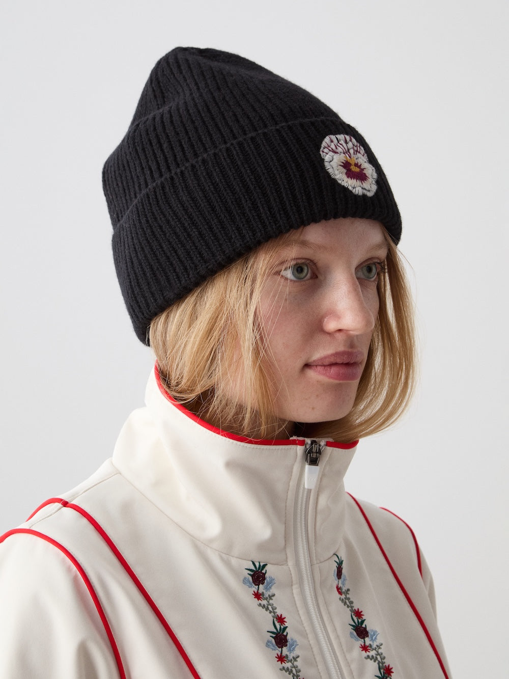 A blonde person wears the We Norwegians WN x Markarian Hat Women, a limited edition black knit beanie with a round embroidered patch, paired with a white zip-up jacket with red piping and floral embroidery, looking off to the side.