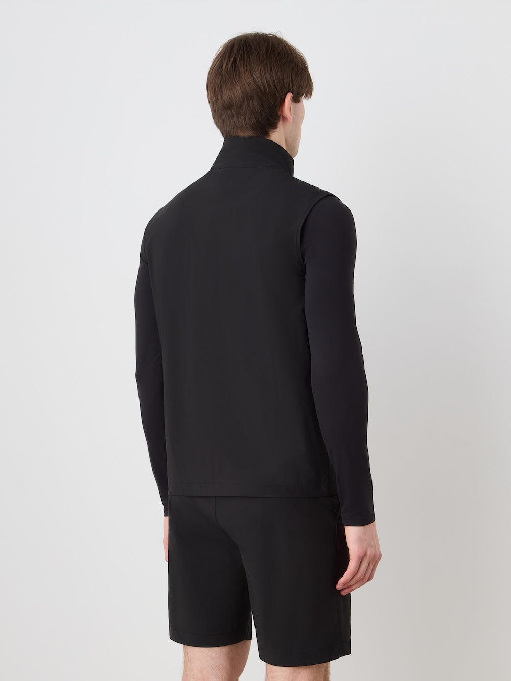 A man with brown hair, seen from behind, models the We Norwegians Albatross Vest Men over a long-sleeve black shirt and black shorts, standing against a plain white background.