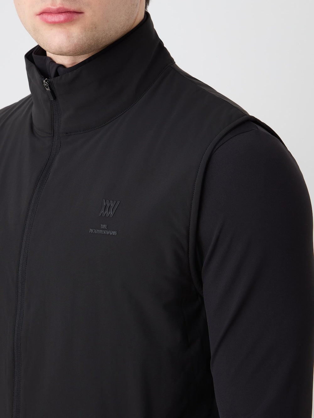 A man is shown from shoulders to mid-chest wearing a black We Norwegians Albatross Vest Men over a black long-sleeve shirt. The golf vest has a small embroidered logo on the left side. The background is light and plain.