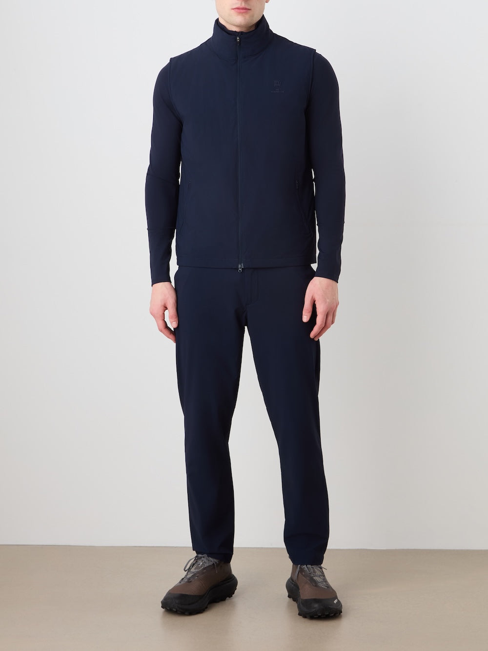 A person stands indoors against a plain white wall, wearing the We Norwegians Albatross Vest Men in dark navy over a long-sleeve shirt, paired with matching navy pants and dark sneakers.