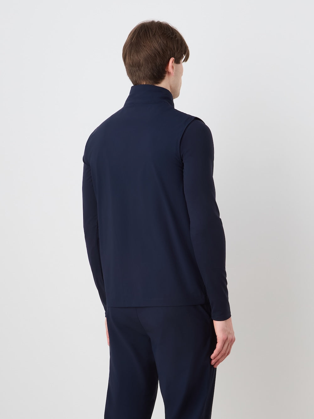 A person with short brown hair stands facing away from the camera, wearing the We Norwegians Albatross Vest Men over a long-sleeve navy blue jacket and matching pants, set against a plain white background.