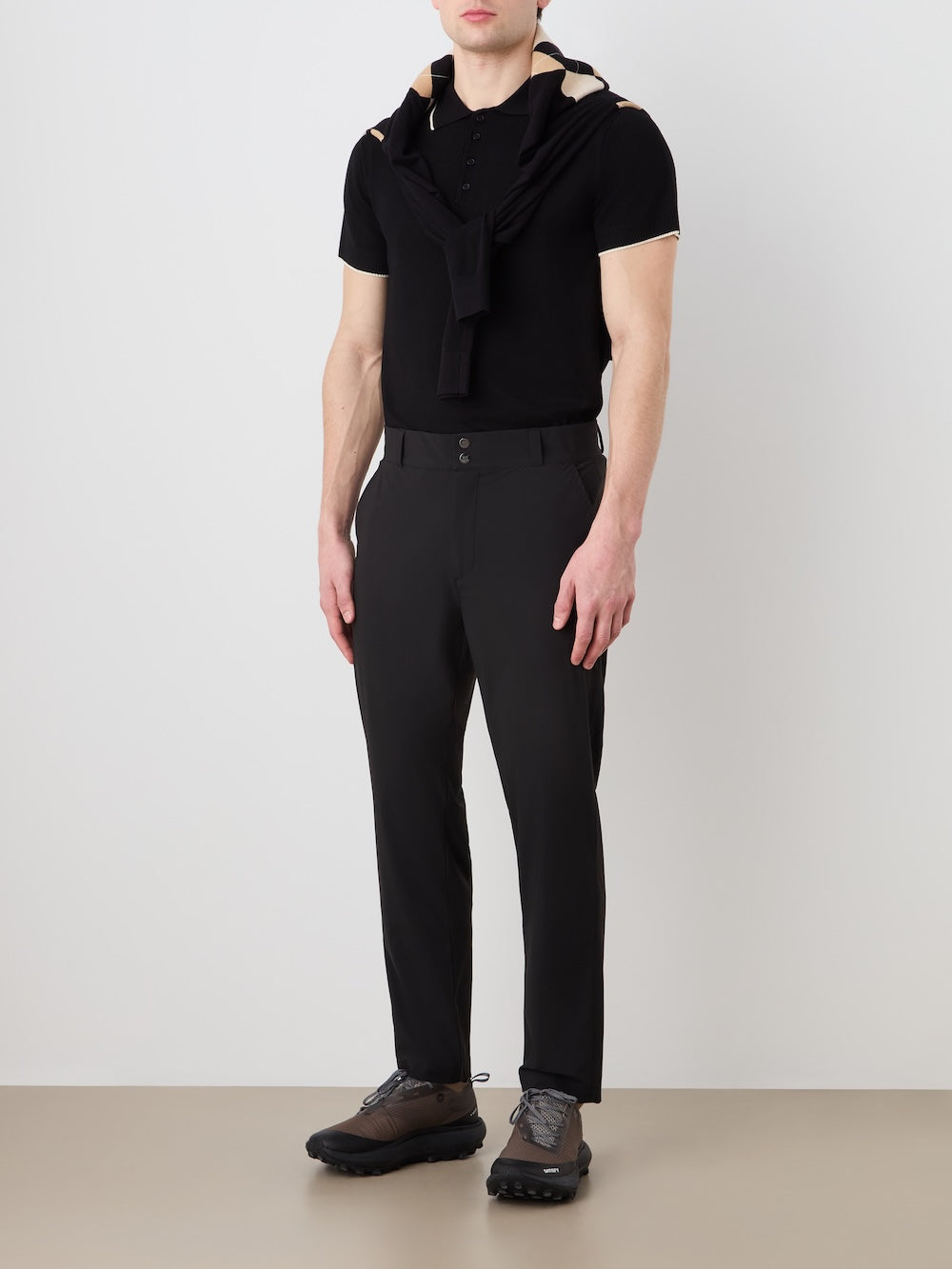 A man stands against a plain background wearing a black polo, dark sneakers, a black sweater over his shoulders, and We Norwegians Albatross Pant Men—breathable, 4-way stretch men's golf pants. His face is partially out of frame.