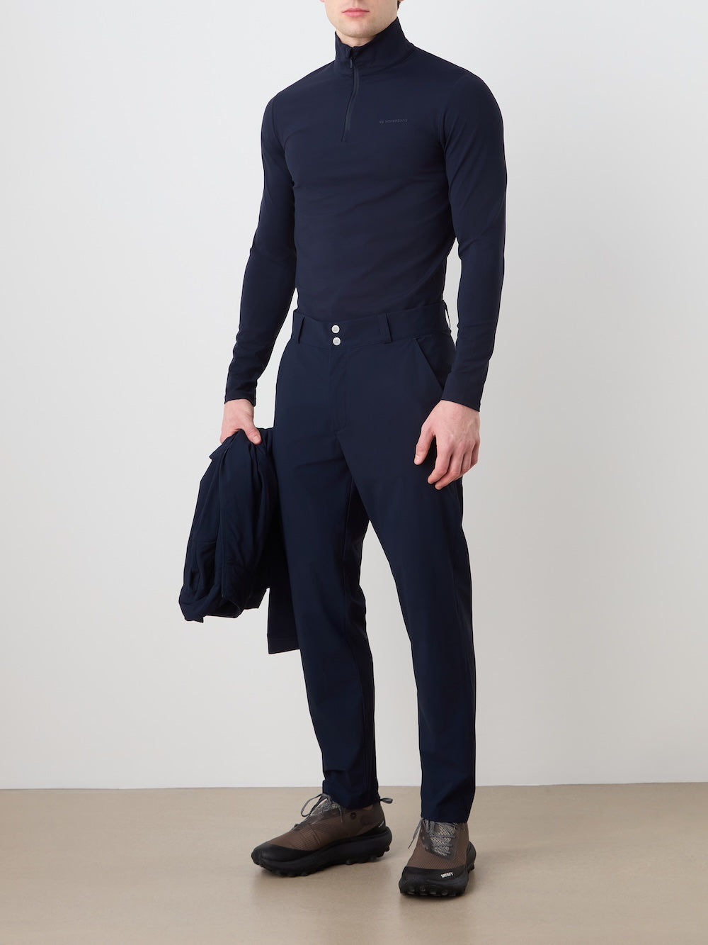 A man stands against a plain wall wearing a fitted, long-sleeve navy half-zip top and We Norwegians Albatross Pant Men with 4-way stretch. He holds a jacket in his left hand, wears dark sneakers, and his face is not visible.