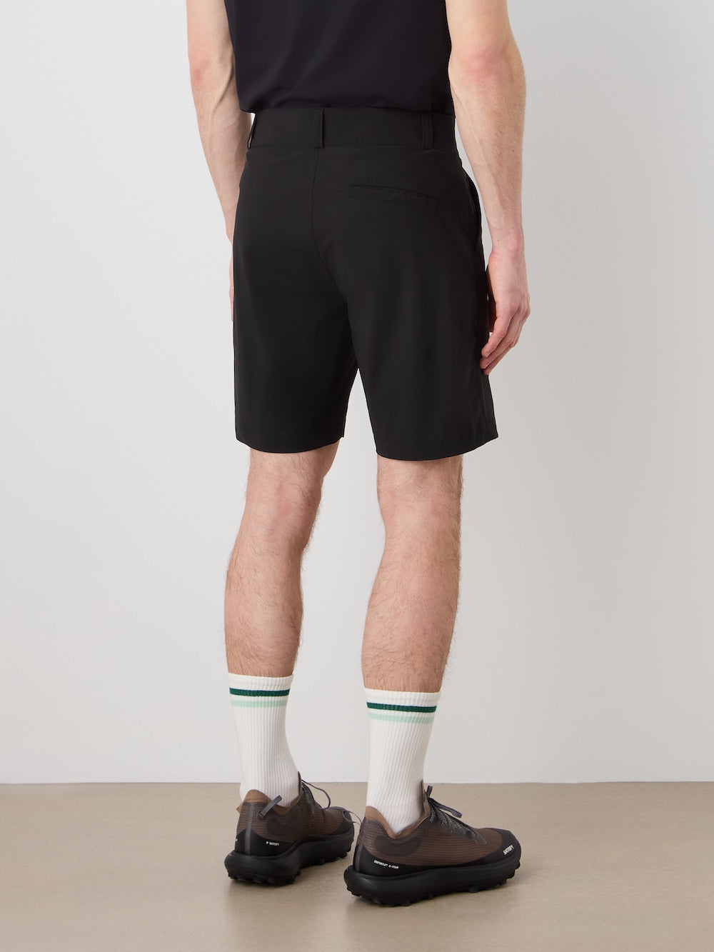 A person faces away, wearing a black shirt, We Norwegians Albatross Shorts Men, white socks with green stripes, and brown athletic shoes against a plain light-colored wall and floor.