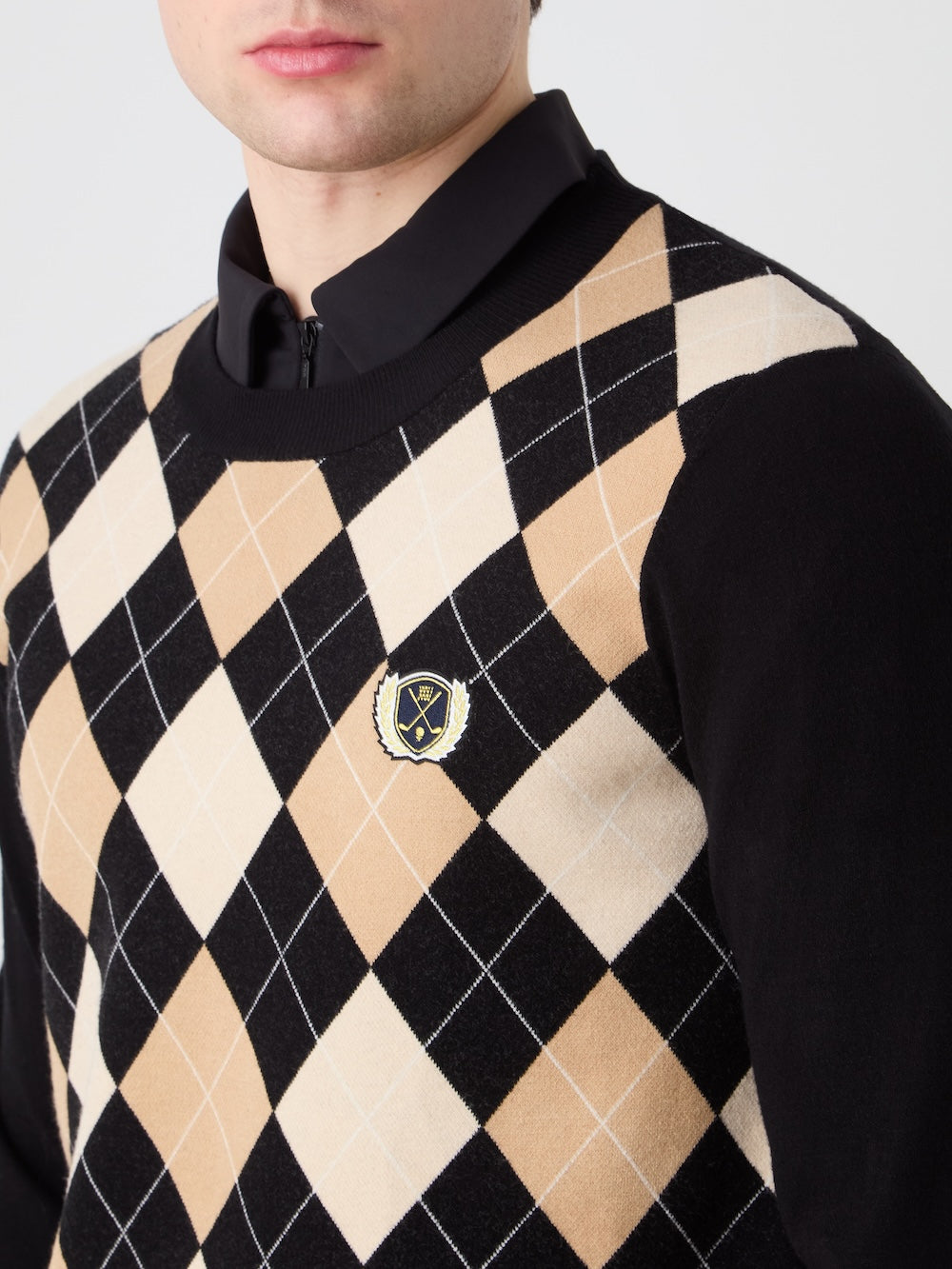 A man wears the We Norwegians Fairway Sweater Men—a beige, black, and white argyle knit with a small crossed golf clubs crest—over a black collared shirt. This lightweight, breathable golf sweater partly obscures his face.