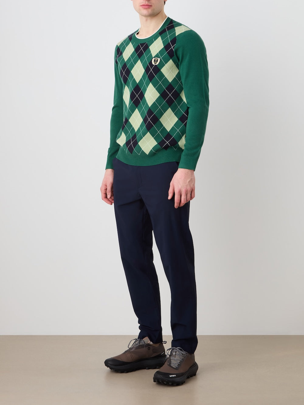 A person stands against a plain wall wearing the We Norwegians Fairway Sweater Men, a green argyle-knit, lightweight and breathable sweater with navy and light yellow diamonds and a small chest crest, paired with navy trousers and dark athletic shoes.