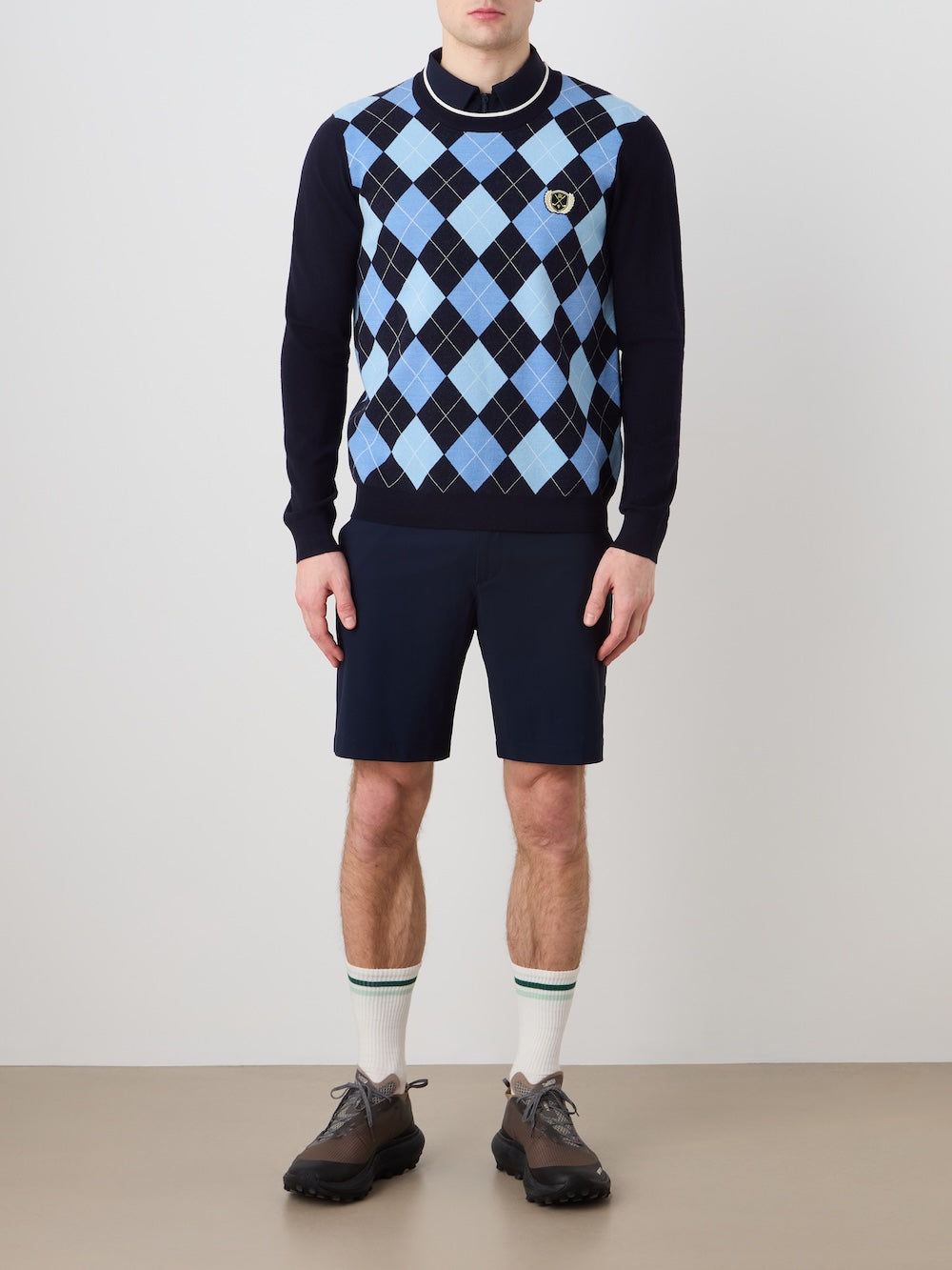 A man models the We Norwegians Fairway Sweater Men against a plain background, pairing the blue and black argyle knit with navy shorts, green-striped white socks, and dark sneakers. His hands are at his sides; his face isn't shown.