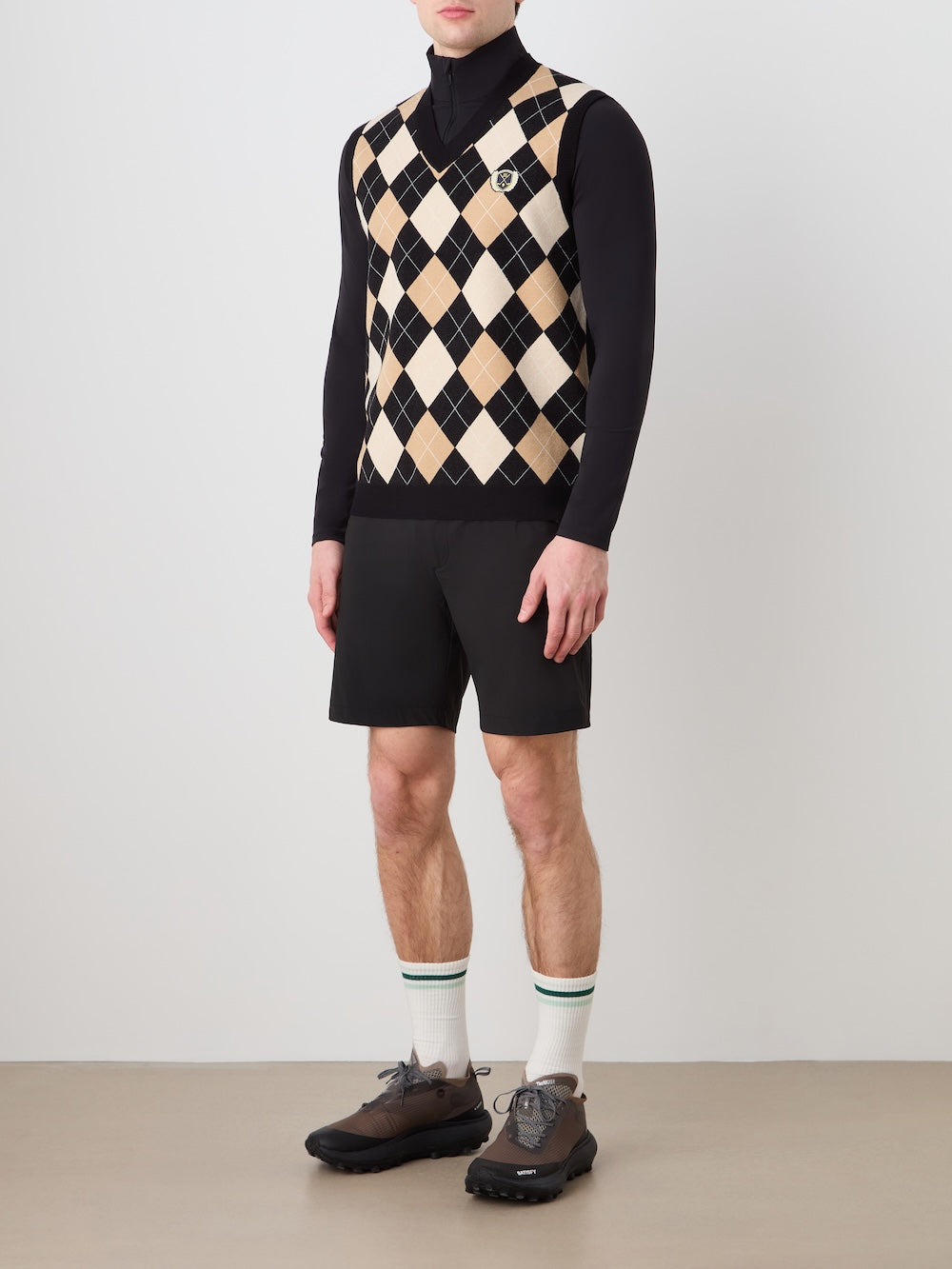 A man models the We Norwegians Fairway Vest Men over a black turtleneck, paired with black shorts, white socks with green stripes, and gray sneakers against a plain background. His head is not shown.