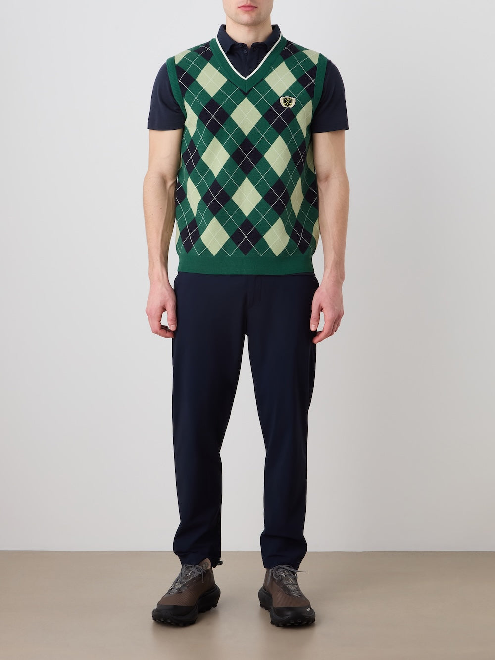 A person models the We Norwegians Fairway Vest Men—a green and navy argyle knit golf vest—over a dark polo, with dark trousers and brown shoes. Their face is not shown; hands are at their sides against a plain background.