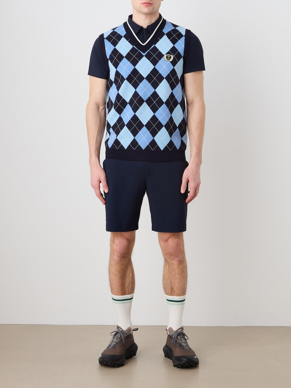 A man stands against a plain background wearing the We Norwegians Fairway Vest Men over a navy polo, paired with navy shorts, white socks with green stripes, and black sneakers. His arms are by his sides and his face is not visible.