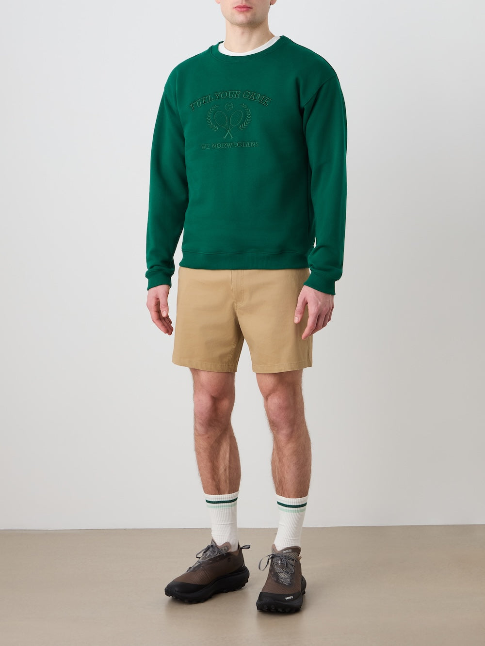 A person stands against a plain wall wearing the We Norwegians Bounce Sweater Men in green, tan shorts, white socks with green stripes, and dark sneakers. Their head is not fully visible in the image.