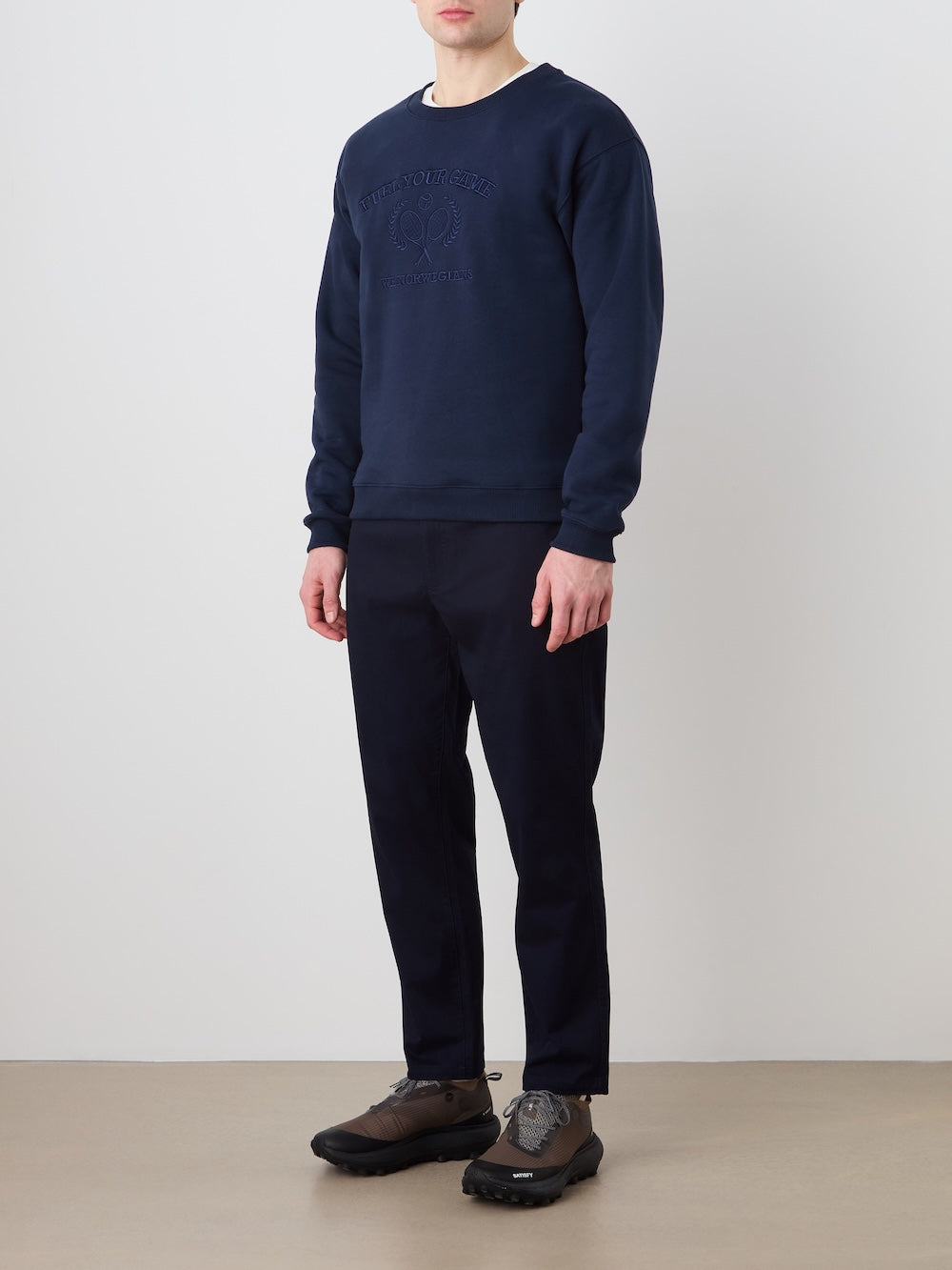 A man stands against a plain, light wall wearing the We Norwegians Bounce Sweater Men in navy and dark navy pants with brown sneakers. His relaxed pose and the soft sweater evoke a subtle vintage tennis style.