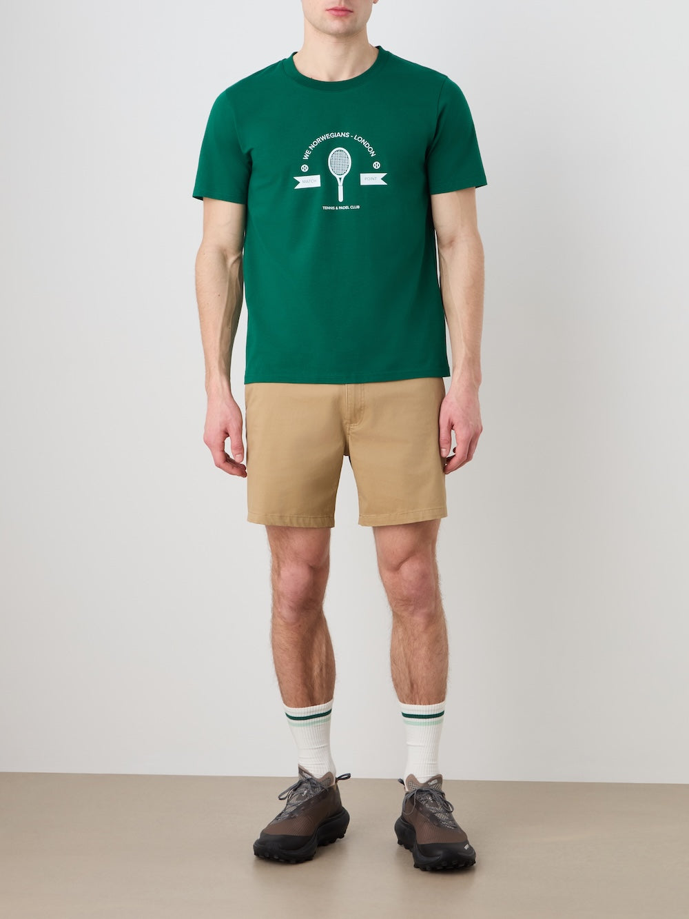 A person stands against a plain background wearing the We Norwegians Love Tee Men, featuring a vintage tennis poster graphic, paired with tan shorts, green-striped white socks, and dark sneakers. Their face is not visible.