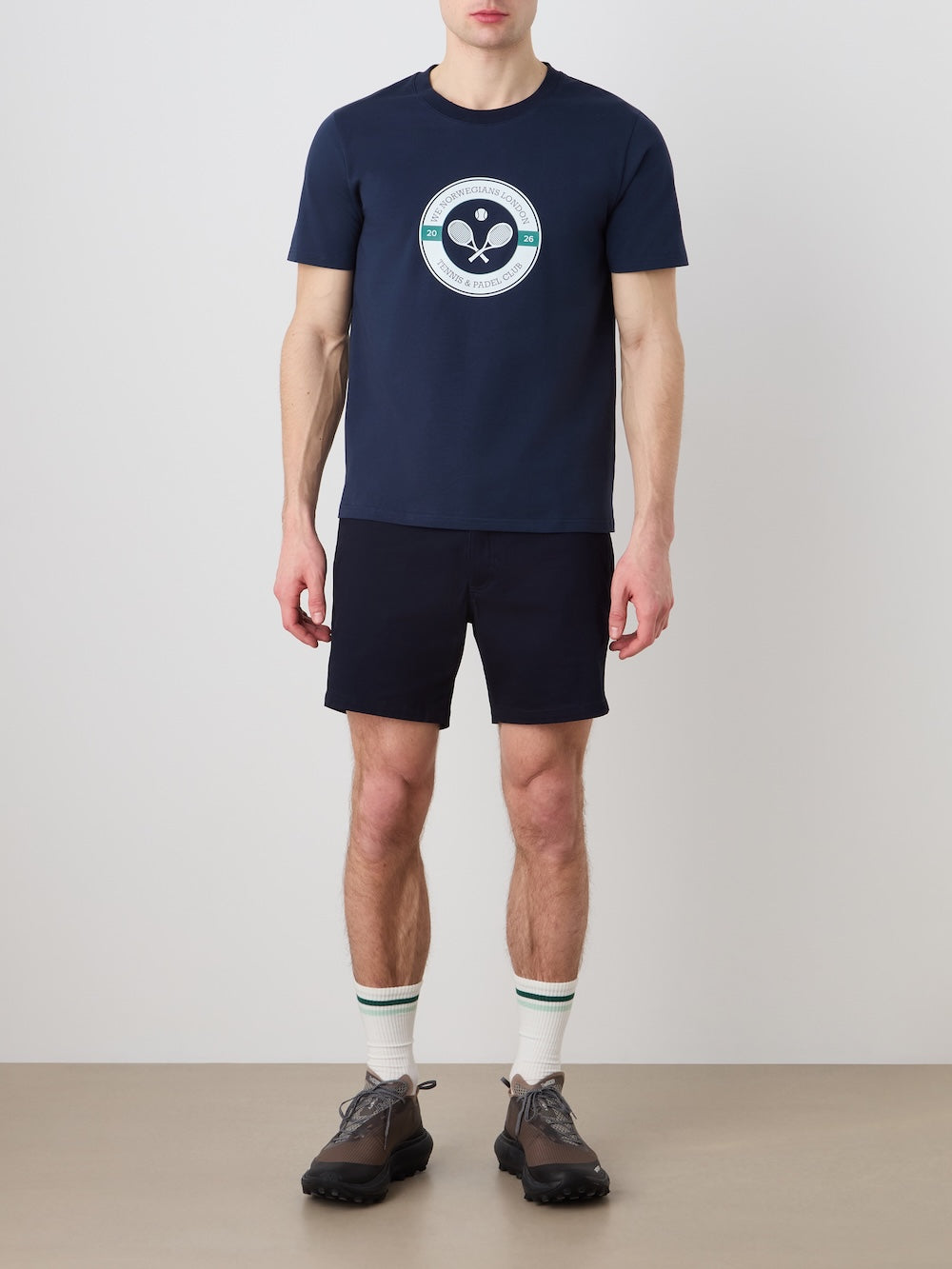 A man poses against a plain background wearing the We Norwegians Love Tee Men—a navy blue cotton T-shirt with a retro tennis-inspired round logo—navy shorts, white socks with green stripes, and dark athletic shoes.