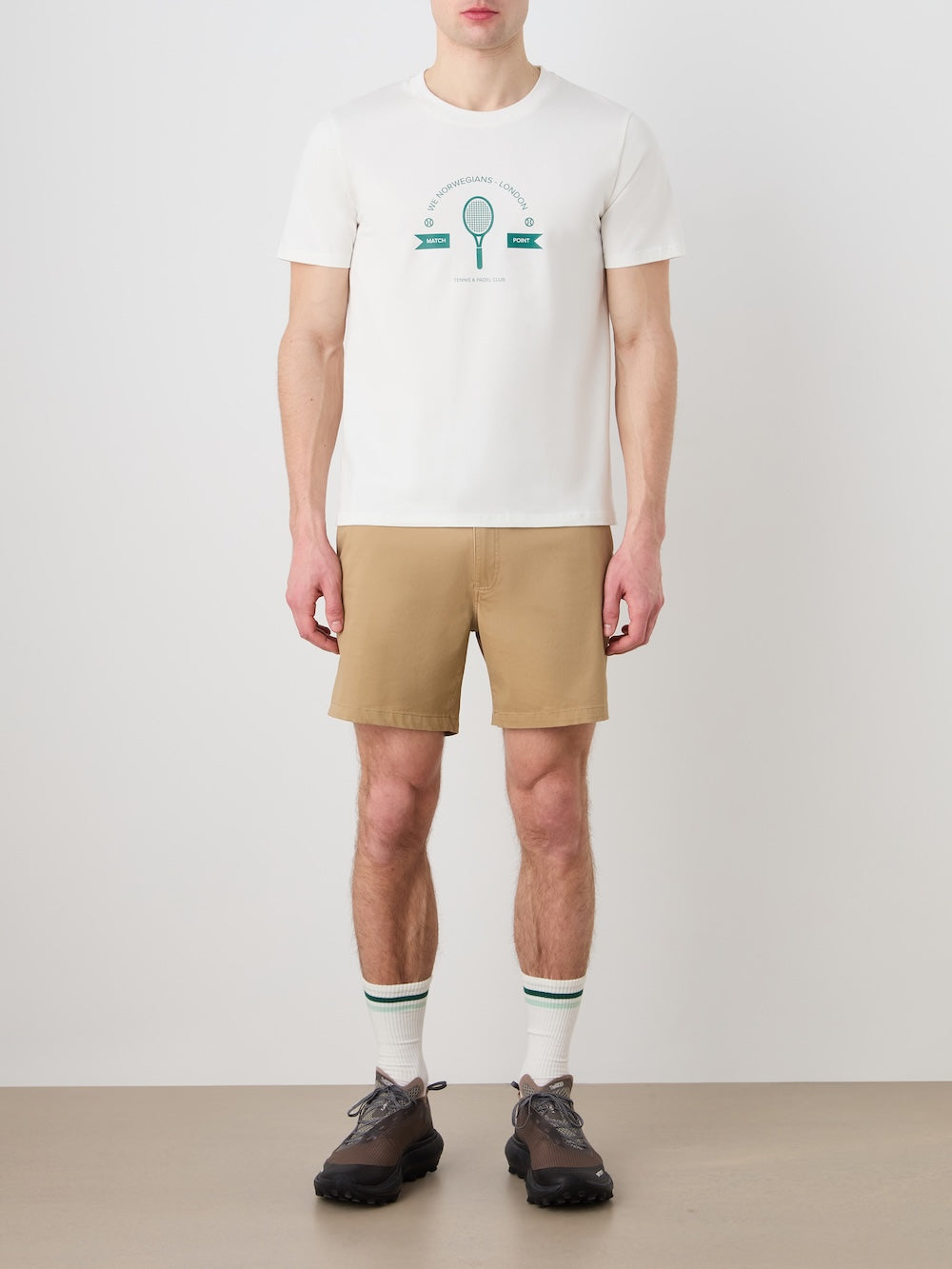 A man models the We Norwegians Love Tee Men against a plain backdrop, paired with tan shorts, retro-striped socks, and black sneakers. His relaxed pose channels vintage tennis poster vibes; his face is partially out of view.