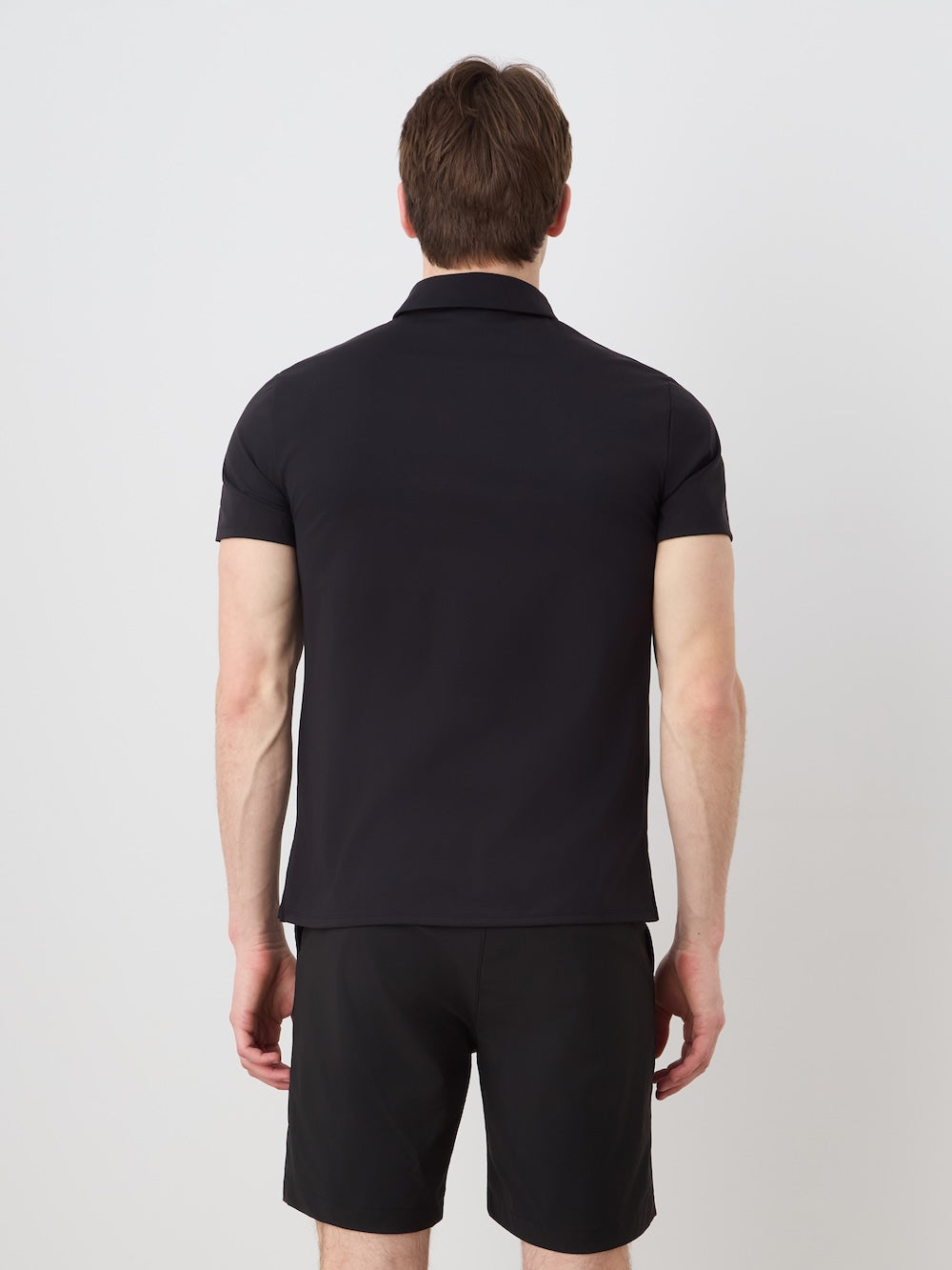 A person with light skin and brown hair faces away from the camera, wearing the We Norwegians Honor Zipped Polo Men and black high-performance shorts, set against a plain white background.