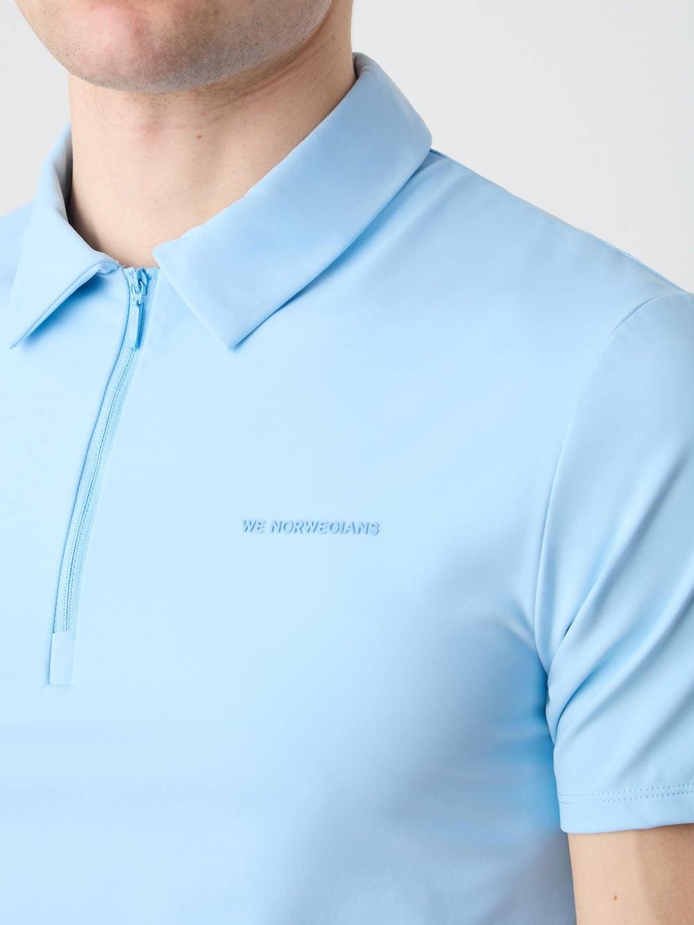 A man wears the light blue Honor Zipped Polo Men by We Norwegians, featuring a chest zipper and subtle "WE NORWEGIANS" branding. The cropped image highlights the upper torso and showcases its high-performance fabric.