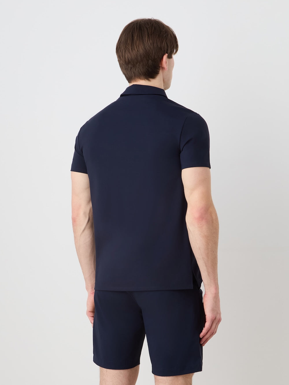 A man with short brown hair faces away from the camera, wearing We Norwegians' Honor Zipped Polo Men in dark navy and matching shorts, made from high-performance fabric, against a plain light background.