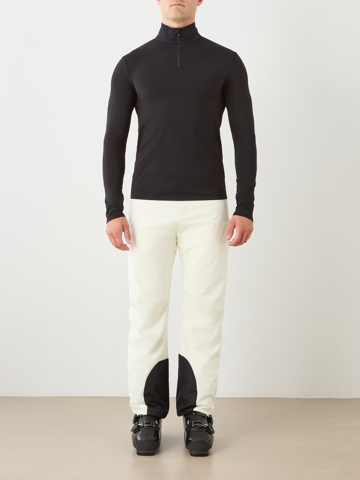 A person stands facing forward against a plain background, wearing the We Norwegians Honor Zipped Long Sleeve Men top in black, paired with white pants featuring black patches at the ankles and black ski boots.