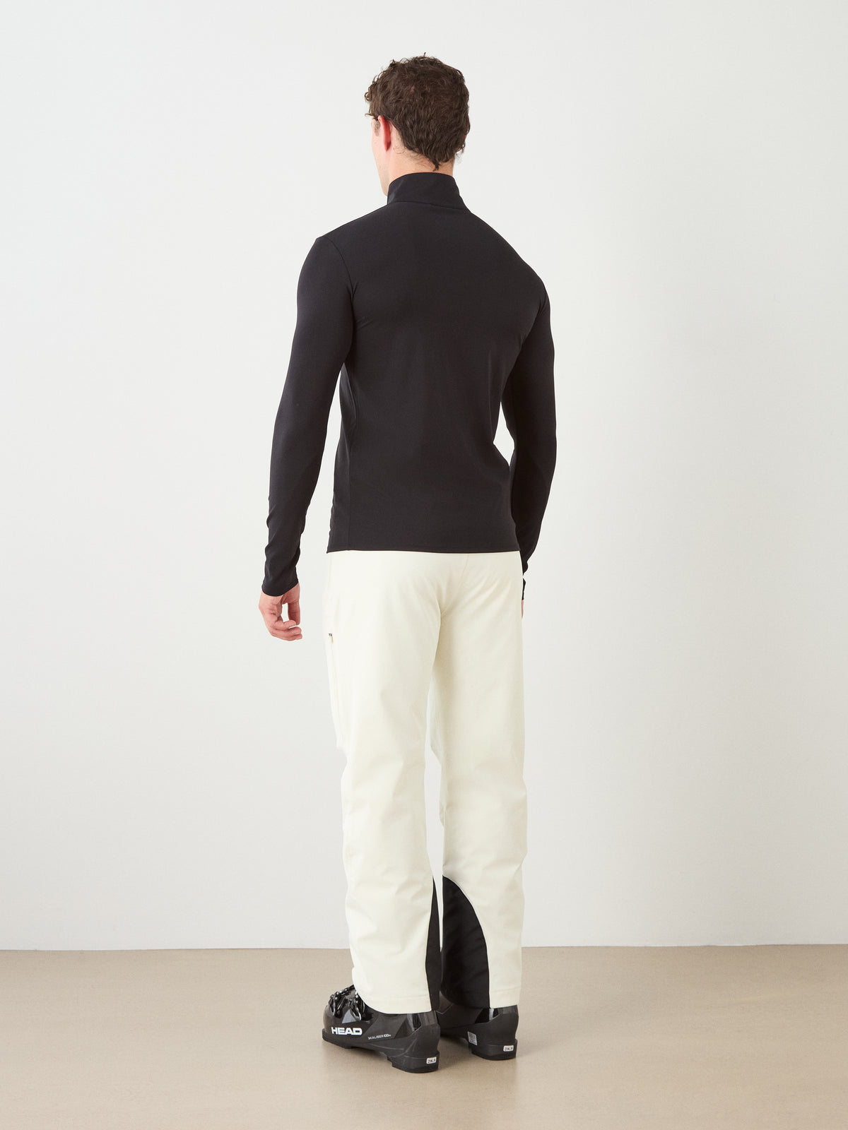 A person stands facing away, wearing the We Norwegians Honor Zipped Long Sleeve Men—a fitted black long-sleeved t-shirt made from high-performance, moisture-wicking fabric—paired with white pants and black shoes, set against a plain light wall and tan floor.