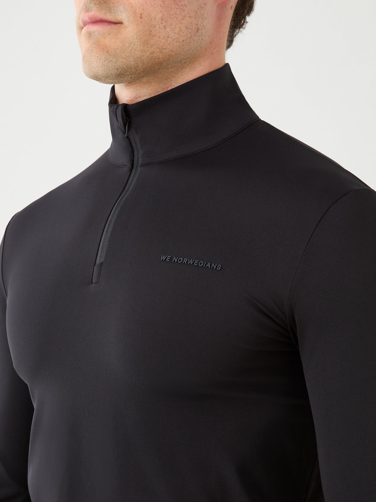 A person wearing the We Norwegians Honor Zipped Long Sleeve Men top in black, featuring a zip-neck design and high-performance fabric, with “WE NORWEGIANS” printed on the chest. The image is cropped to display only the upper torso and lower face.