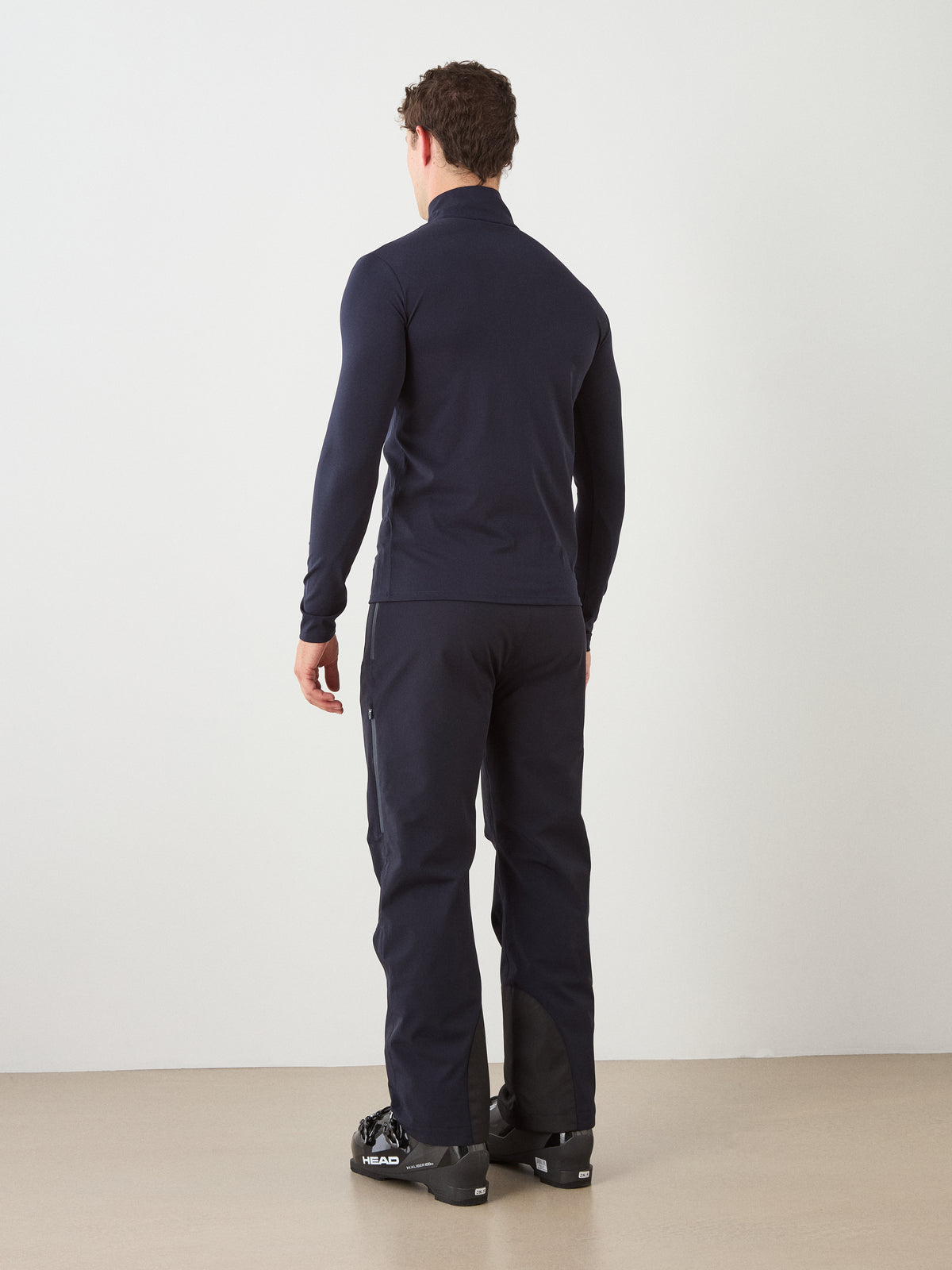A person stands indoors facing away, wearing the We Norwegians Honor Zipped Long Sleeve Men jacket in navy, paired with matching pants and black ski boots. The background features a plain white wall and a beige floor.