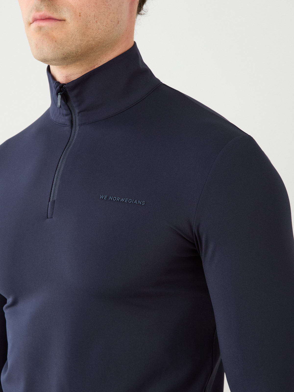 A man is shown from the shoulders to the mouth against a plain background, wearing the Honor Zipped Long Sleeve Men—a fitted navy blue zip-up top by We Norwegians, featuring high-performance fabric and “WE NORWEGIANS” written on the chest.