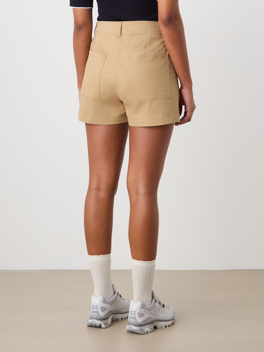 A person stands indoors facing away, wearing a black top and tan We Norwegians Sol Shorts Women—ideal summer shorts for women—paired with white socks and light gray sneakers, against a plain neutral background.