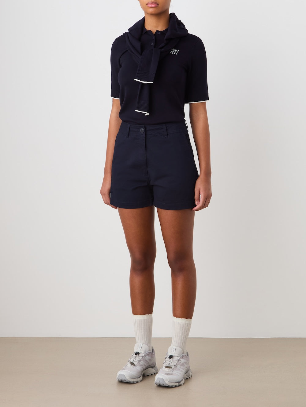 A person stands against a plain background wearing a dark short-sleeve shirt, a sweater tied around their shoulders, We Norwegians Sol Shorts for women, white calf socks, and light gray sneakers.