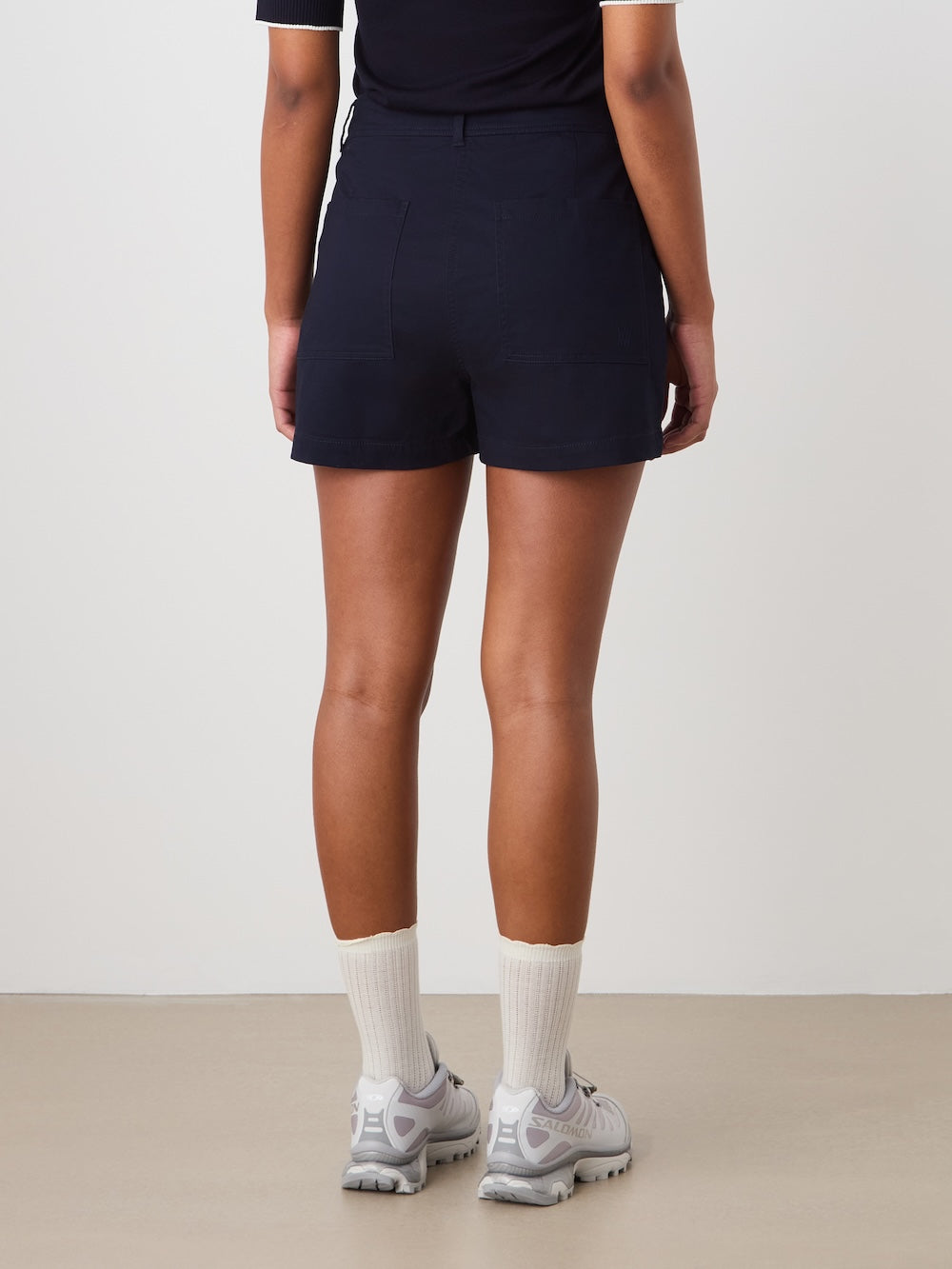 A person stands facing away, wearing a black t-shirt and We Norwegians Sol Shorts Women—dark high-waisted cotton-blend shorts—with white socks and gray sneakers, set against a plain light background.