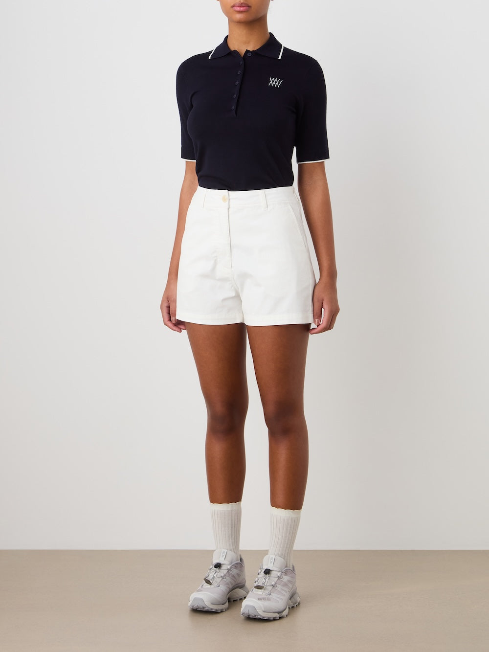 A person wears a black short-sleeve polo, We Norwegians Sol Shorts for women in a crisp cotton-blend, white crew socks, and gray sneakers while standing against a plain light wall. Their face is not visible.