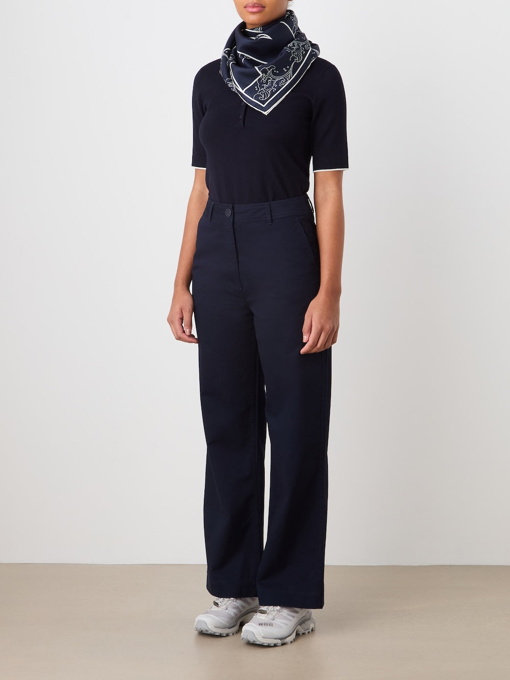 A person, face not visible, stands against a plain background wearing We Norwegians Sol Pant Women sailor trousers, a dark cotton-blend short-sleeve top, light gray sneakers, and a navy patterned scarf tied around their neck.