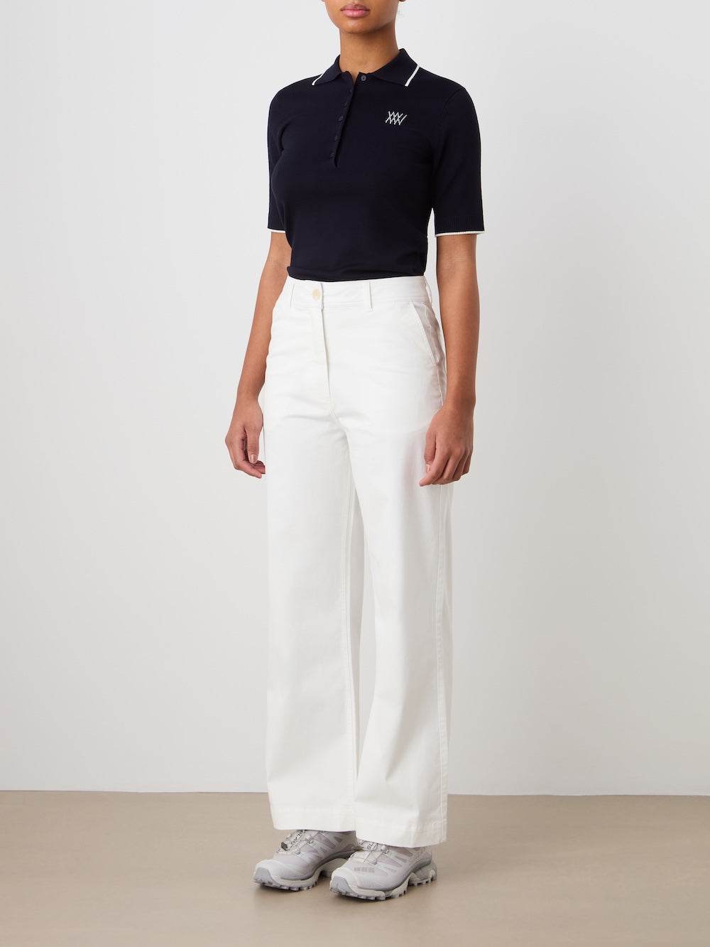 A person stands against a plain background wearing a black short-sleeve polo, We Norwegians Sol Pant Women in high-waisted vintage white, and gray sneakers. Their arms are relaxed at their sides.