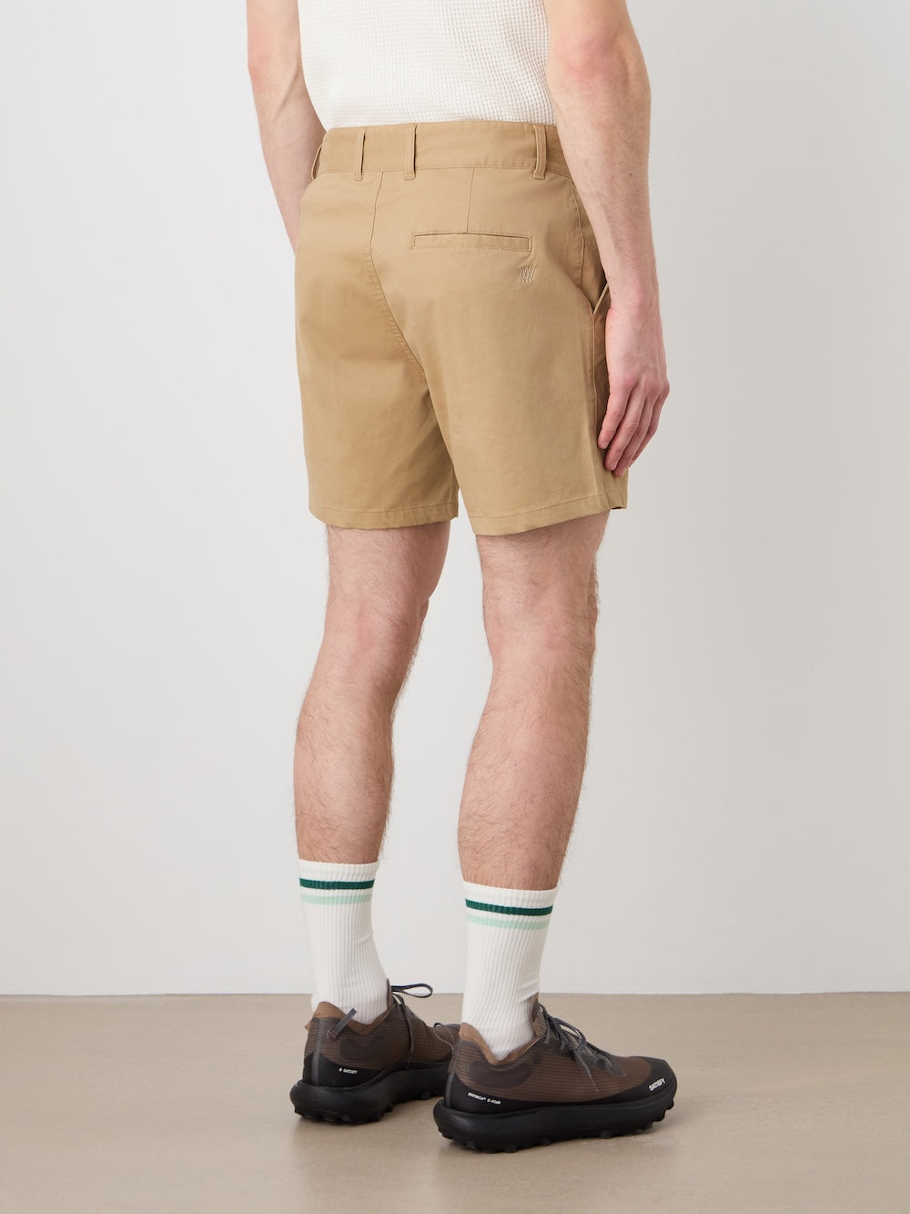 A person wearing We Norwegians Sol Shorts Men, a white ribbed tank top, white socks with green stripes, and dark sneakers stands facing away from the camera against a plain background.