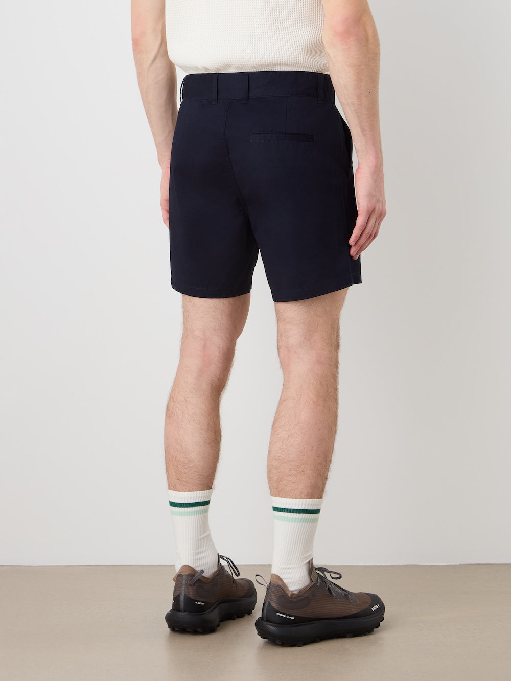 A person stands indoors facing away, wearing a white short-sleeve shirt and We Norwegians Sol Shorts Men in navy, paired with white socks with green stripes and brown shoes. The background is plain and minimal.