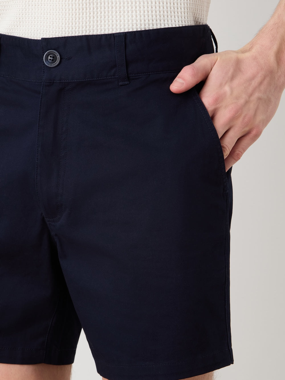 A person wearing We Norwegians Sol Shorts Men in dark navy and a white textured top stands with one hand in their pocket, showing only the lower torso and upper legs.