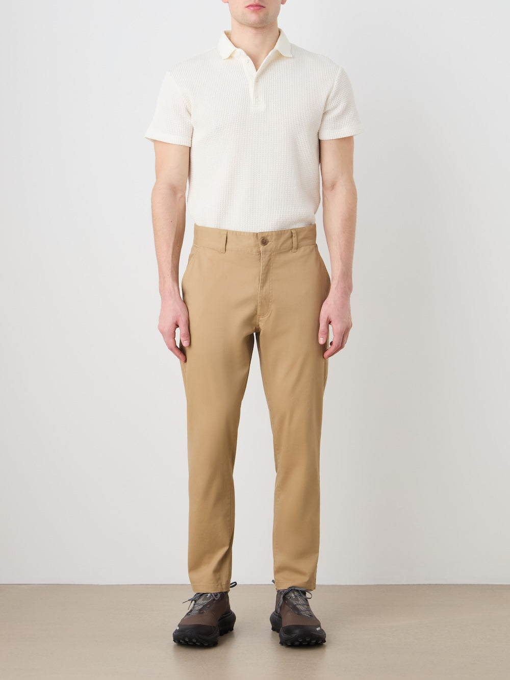 A person stands against a plain white wall wearing a short-sleeve cream polo, We Norwegians Sol Pant Men tan chinos, and black hiking shoes, with arms relaxed at their sides.